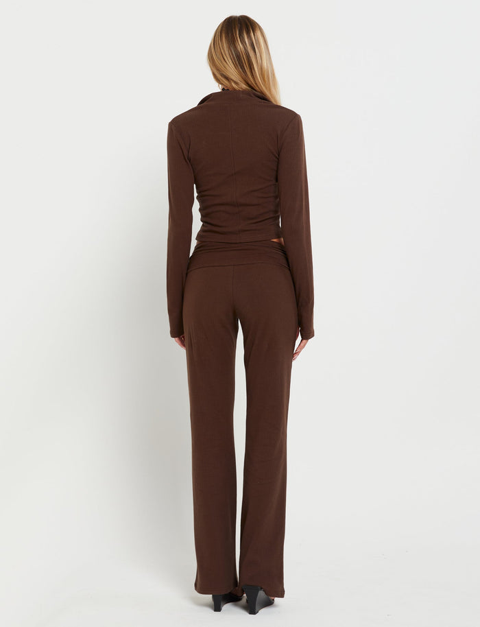 A person with long blonde hair, dressed in the TIGERMIST KAVAT PANT - BROWN and a matching fitted brown long-sleeve top, stands facing away from the camera against a white background. The outfit is simple and elegant, with the person standing in a relaxed posture.
