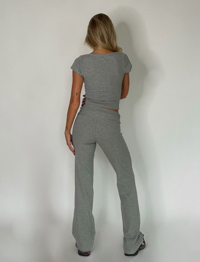 A person with long blonde hair is standing and facing a white wall, wearing a TIGERMIST tight-fitting, short-sleeved grey top and matching Dove Grey KAVAT pants. They stand confidently with one hand on their hip and the other arm relaxed by their side.