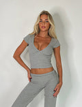 A woman with long blonde hair is wearing a fitted TIGERMIST KAVAT TOP - GREY : DOVE GREY crop top and matching grey pants. She stands against a plain white background with one hand on her hip and the other relaxed by her side.