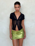 A person stands against a plain light-colored wall, flaunting a black sheer short-sleeved top with a deep neckline and a small black collar. They're styled in the Kaye Skirt from TIGERMIST, featuring green sequins, and large hoop earrings, their hair neatly arranged.