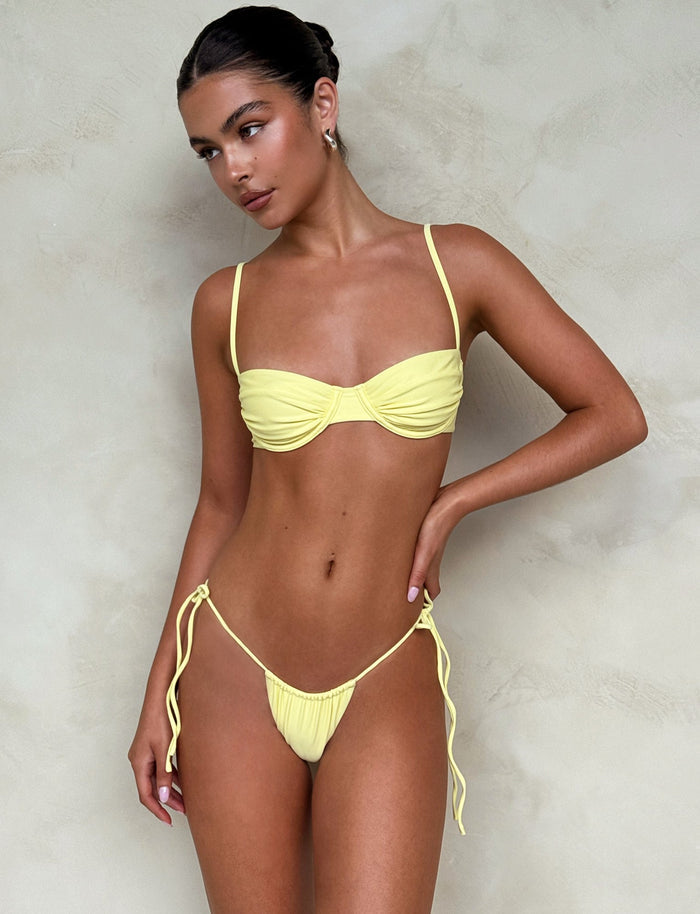 A woman stands against a light gray textured background wearing the TIGERMIST KHESSA Bikini Top in Lemon yellow, featuring tied side strings on the bottom. Her hair is pulled back, and she looks to the side with a neutral expression, accessorized with chic earrings.