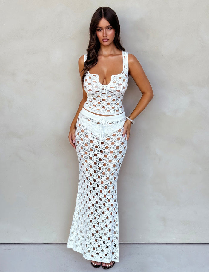 A woman with long, dark hair is standing against a plain, light-colored wall. She is wearing a white crochet two-piece outfit from TIGERMIST, featuring the Kincade Skirt - White and a sleeveless crop top. She has a bracelet on her right wrist and is looking directly at the camera.