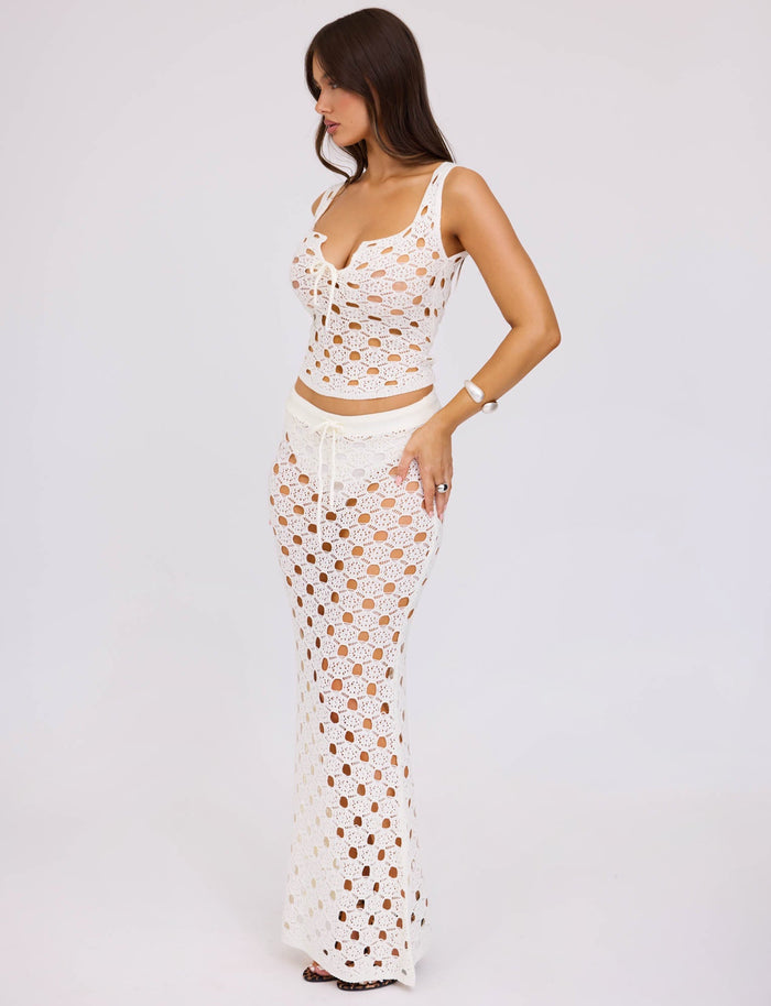 A woman with long brown hair is wearing the KINCADE TOP - WHITE from TIGERMIST. The top has an intricate crocheted design exposing her skin underneath. She stands against a plain white background, looking to her right with one hand on her hip.