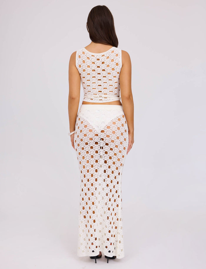 A woman stands facing away, wearing the Kincade Top in white from TIGERMIST, paired with a matching long skirt. The outfit features an intricate see-through crochet pattern. She has long brown hair and is wearing black high-heeled shoes against a plain, light-colored background.