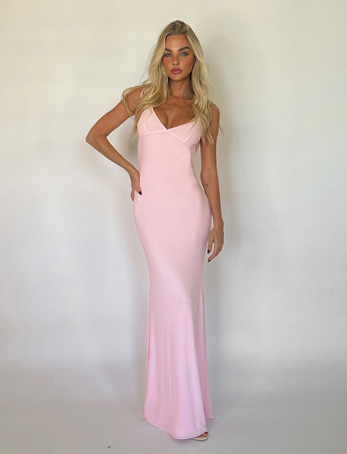A woman with long blonde hair stands against a plain white background, wearing a floor-length, sleeveless KYLAH DRESS in baby pink by TIGERMIST. She is posing with one hand on her hip and the other hanging loosely by her side.