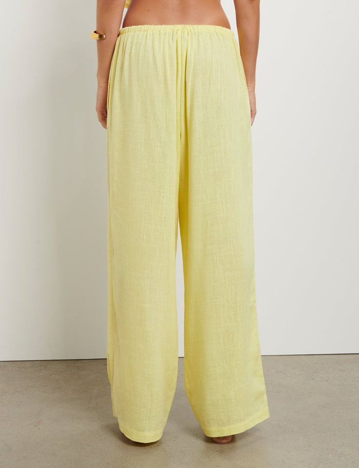 A person is shown from the back, wearing loose-fitting, high-waisted LAINEY PANT - YELLOW : LEMON pants by TIGERMIST. The person is barefoot and standing on a smooth, light-colored surface in front of a plain white wall.