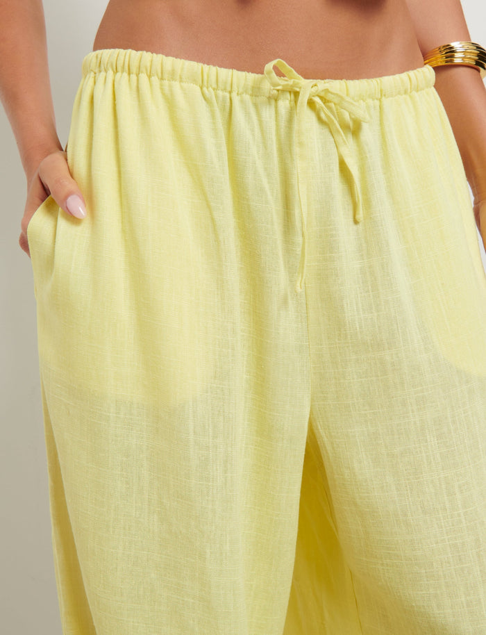 The image shows a close-up of a person wearing the LAINEY PANT - YELLOW: LEMON by TIGERMIST. These high-waisted, loose-fitting pants feature an elastic waistband with a drawstring. The person's hands are tucked into the front pockets, and a golden bracelet is visible on their right wrist.