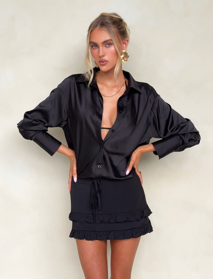 A woman with blonde hair stands against a light background, wearing a partially open black satin button-up shirt from Tiger Mist that reveals a bralette underneath. She completes her outfit with the LEOLA SKIRT - BLACK by TIGERMIST, which features a ruffled hem detail, and she's accessorized with gold jewelry.