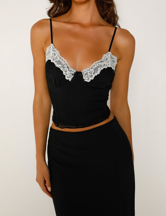 A woman wearing a LEYLA TOP - BLACK by TIGERMIST, with thin straps and a lace trim along the top edge, paired with a matching black skirt. The image is cropped from the shoulders down, highlighting her stylish outfit against a plain white background.