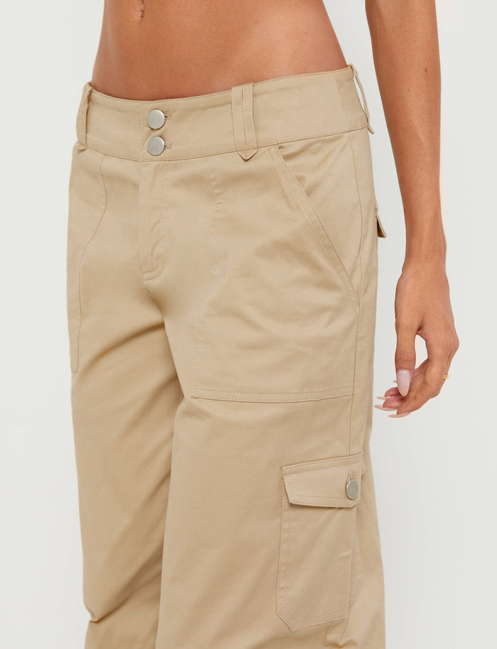 A person is wearing the LISSA PANT - NEUTRAL : BEIGE from the TIGERMIST collection, featuring two front buttons, a side pocket with a buttoned flap, and visible stitching details. The person's left hand is partially visible, resting along the side of the pants against a plain white background.