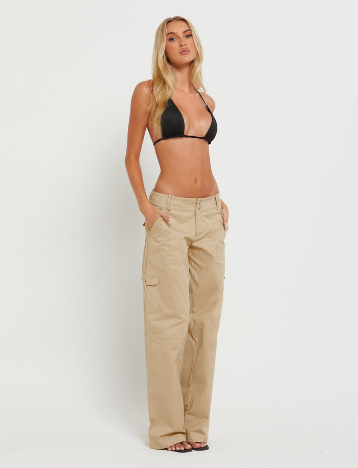 A woman with long blonde hair is standing against a plain white background. She is wearing a black bikini top and the LISSA PANT - NEUTRAL : BEIGE by TIGERMIST, with her hands in the pockets. Her confident facial expression and posture exude self-assurance.