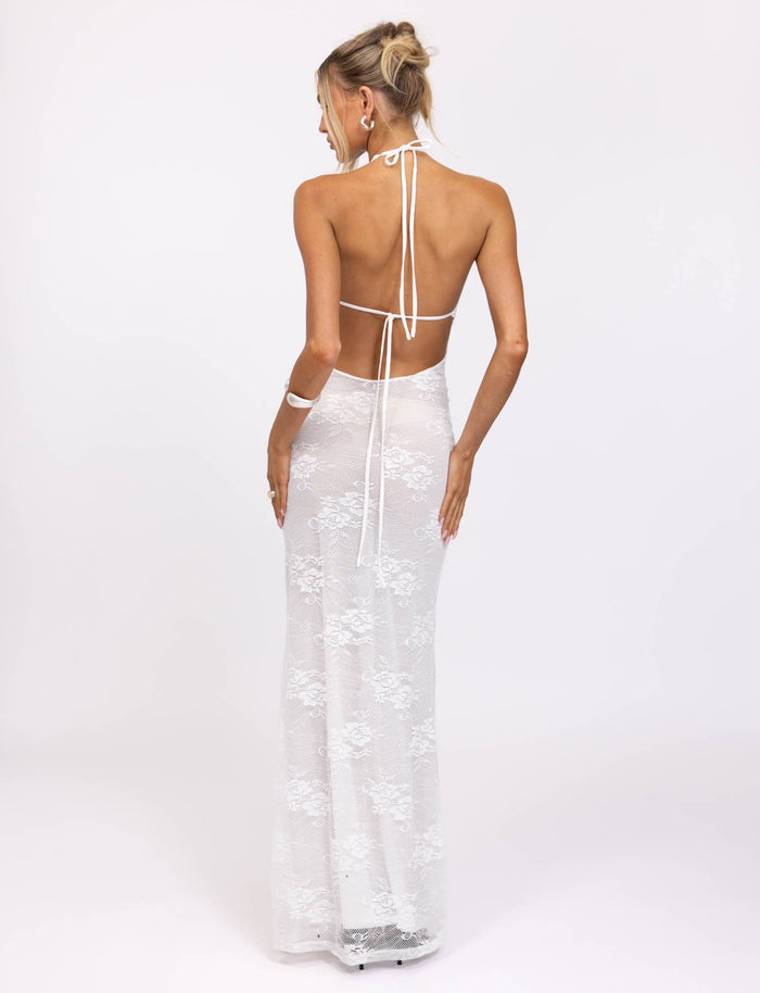 A person is photographed from behind, standing against a plain white background. They are wearing the LUCIA DRESS - WHITE : LACE, a backless halter-style gown by TIGERMIST that extends to the floor. Their hair is styled in an elegant updo.