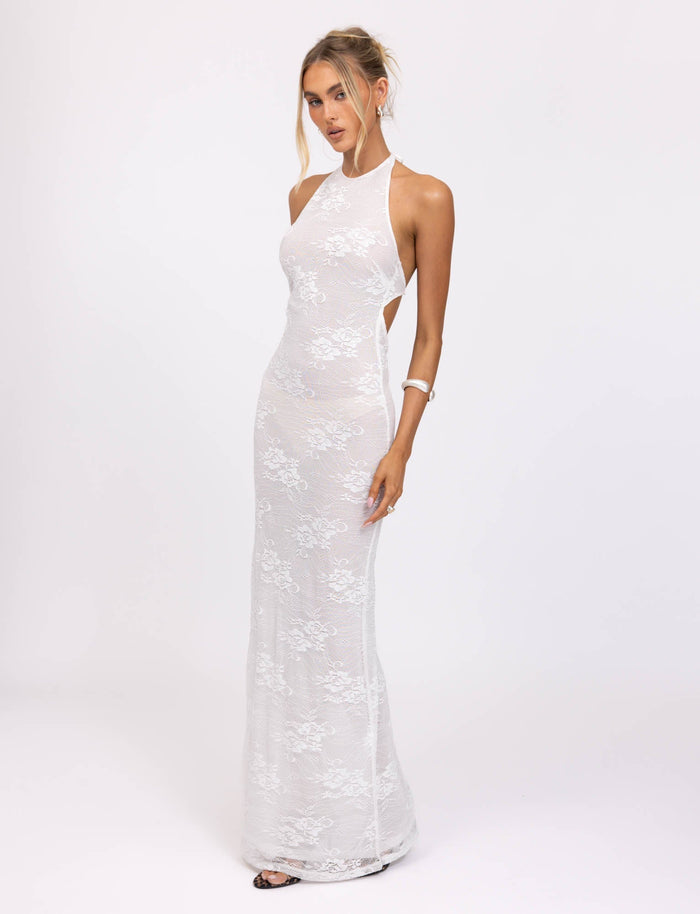 A woman in a fitted, floor-length LUCIA DRESS - WHITE : LACE from TIGERMIST with a halter neckline poses against a plain white background. She has an updo hairstyle and wears minimal jewelry, including a bangle on her right wrist. She appears to be looking slightly downward with a soft expression.