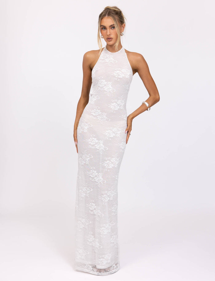 A woman stands against a plain white background, modeling the sophisticated Lucia Dress by Tiger Mist. This elegant white lace gown features a high neckline and a form-fitting silhouette that extends to the floor. She is wearing minimal jewelry, including a bracelet, and her hair is styled up.