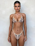 A woman stands against a plain wall, wearing the Lula Bikini Bottom - Brown: Snake by TigerMist. She has her hair tied back and accessorizes with hoop earrings and a small necklace. Her expression is neutral, and her hands are relaxed at her sides.