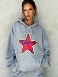 A person wearing the LUMEN HOODIE - GREY from TIGERMIST, featuring a large red star and the text "TIGERMIST" on the front. The person has their hands in the hoodie pockets and is standing against a plain, light-colored wall. They are also sporting matching gray sweatpants proudly showcasing the TIGERMIST logo.