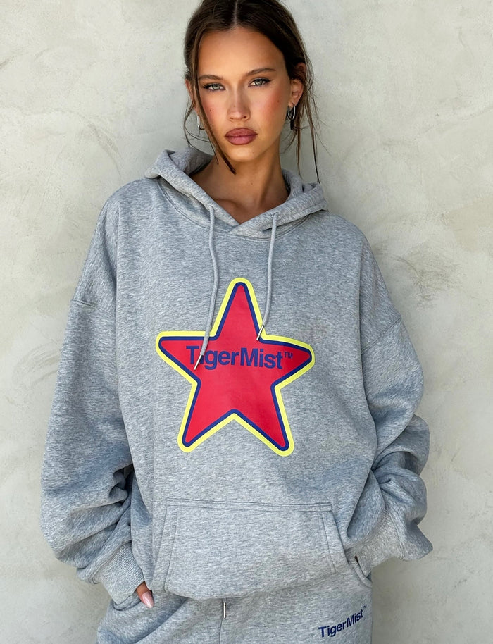 A person wearing the LUMEN HOODIE - GREY from TIGERMIST, featuring a large red star and the text "TIGERMIST" on the front. The person has their hands in the hoodie pockets and is standing against a plain, light-colored wall. They are also sporting matching gray sweatpants proudly showcasing the TIGERMIST logo.