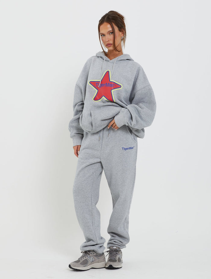 A woman is posing against a white background, donning a light gray LUMEN HOODIE by TIGERMIST with a colorful star graphic, matching gray sweatpants featuring "Tiger Mist" on one leg, and gray athletic shoes. Her hair is tied back, and she has a relaxed stance.