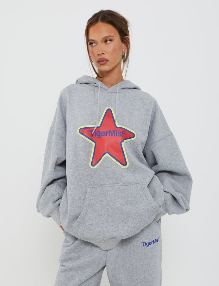 A person wearing the LUMEN HOODIE - GREY from TIGERMIST, featuring a large red star and "TIGERMIST" logo in blue on the front. They have their hands in the hoodie’s pockets and are also sporting matching gray sweatpants with the same "TIGERMIST" logo on the thigh.