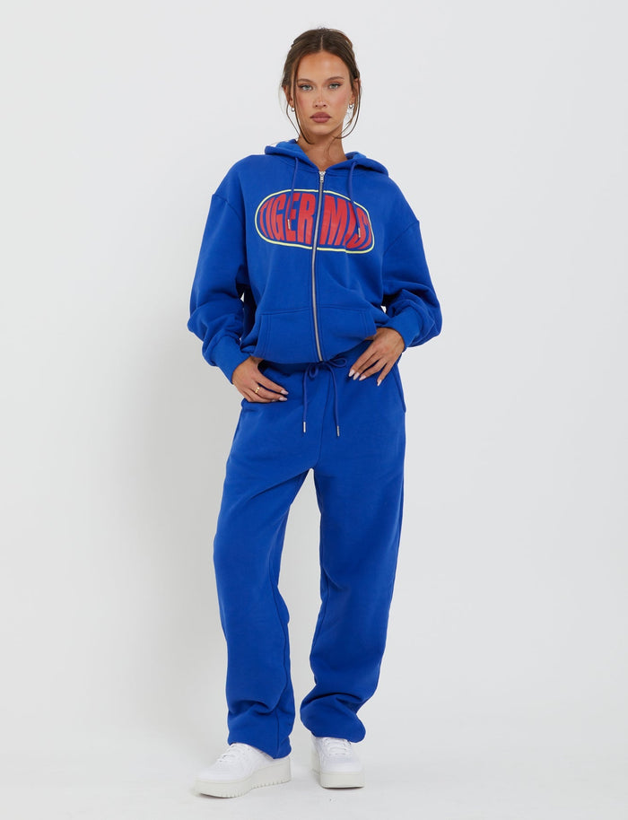 A woman stands against a plain white background, wearing a matching blue tracksuit from Tiger Mist. The hoodie has the word "TIGERMIST" printed on it in red and white letters. She is wearing the LUMEN PANT - BLUE, paired with white sneakers, with her hands in her pockets and looking directly at the camera exuding a Tiger Mist vibe.