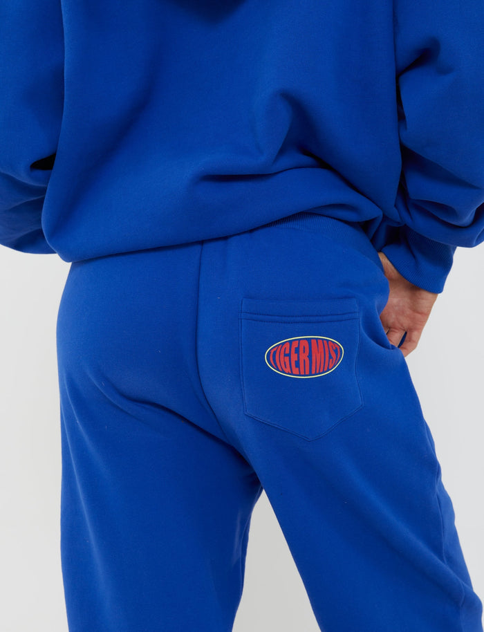 A person dressed in a vibrant blue TIGERMIST sweatshirt and LUMEN PANT - BLUE sweatpants with their back to the camera. The sweatpants feature a small patch on the back pocket that reads "TIGERMIST" in red letters against a blue and red-orange oval background.