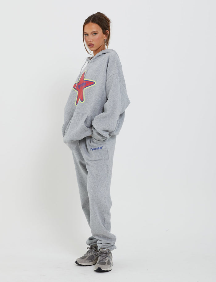 A person stands against a plain white background wearing a grey hoodie from Tiger Mist with a large red star on the chest and matching LUMEN PANT - GREY from TIGERMIST. They have their hands in their pockets and are wearing grey sneakers. Their hair is tied back, and they have a neutral expression.