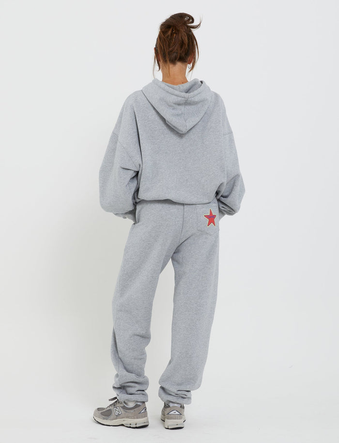 A person with their back to the camera is wearing a loose-fitting grey tracksuit from TigerMist, specifically the Lumen Pant - Grey, along with a matching hoodie. Both pieces are adorned with a small red star on the right pocket. They have brown hair tied up in a messy bun and are standing against a plain white background.