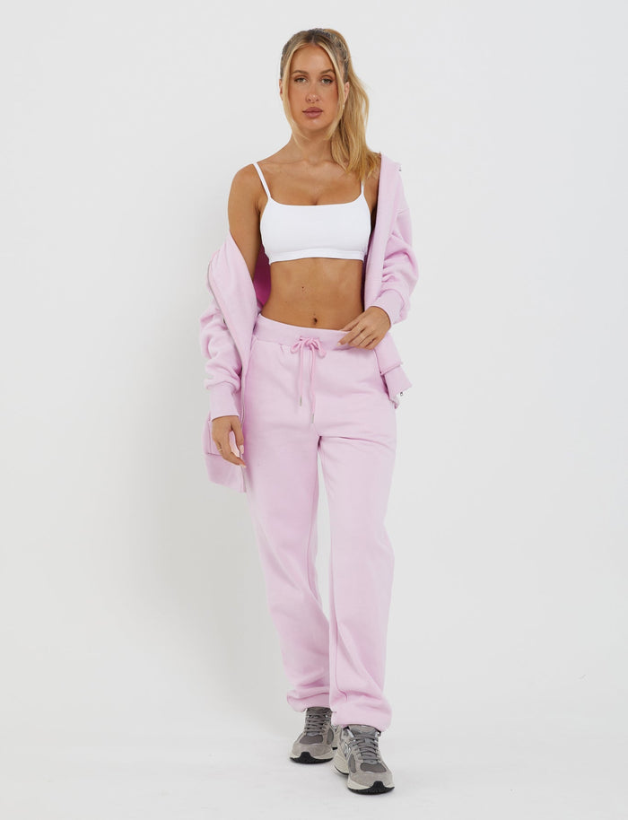 A woman dressed in the TIGERMIST LUMEN PANT - PINK and a white crop top stands against a plain white background. She has her hair tied back and is looking forward, casually holding one side of her open hoodie. The mid-rise waist of the pink LUMEN Pant complements her relaxed look along with gray sneakers.