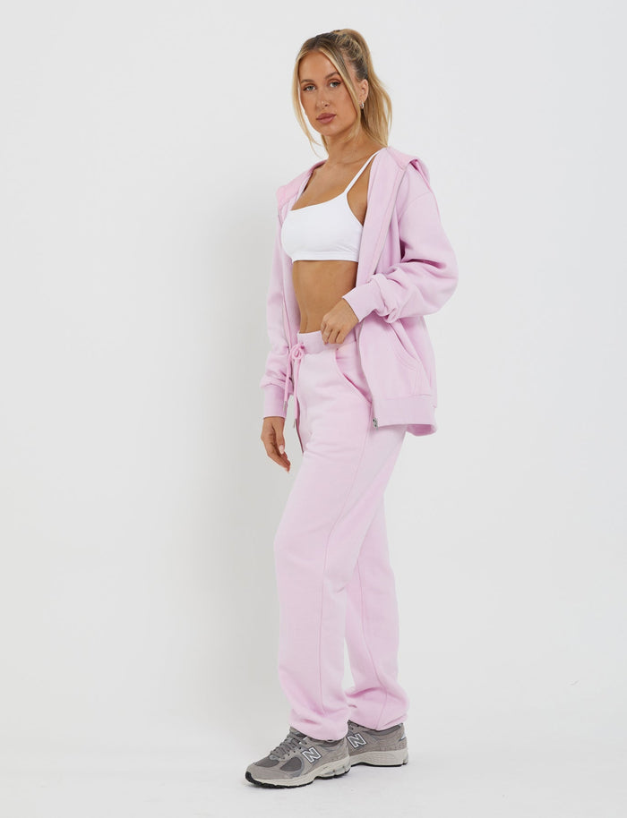 A person with long hair stands against a plain white background, wearing a white sports bra and an oversized pink loungewear hoodie. They pair the outfit with TIGERMIST's LUMEN PANT in pink, mid-rise waist sweatpants, and gray athletic sneakers. With a confident stance and their left hand in their pocket, they exude style and comfort.