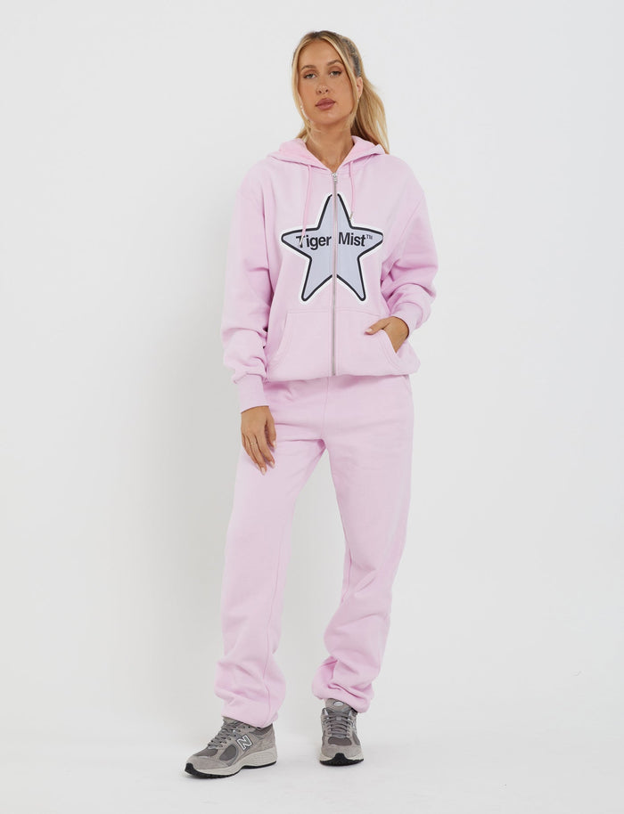 A person stands facing forward, wearing a light pink hoodie with a large star logo and "TIGERMIST" text in the center, paired with the TIGERMIST LUMEN PANT - PINK that features a mid-rise waist in matching light pink. They have their hands in the hoodie pockets and are also wearing gray sneakers. The background is white.