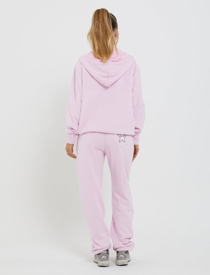A person wearing a light pink hoodie and matching TigerMist LUMEN PANT in pink is standing facing away from the camera. They have long hair tied up in a high ponytail. The joggers feature a small star graphic on the back pocket. The background is plain white.