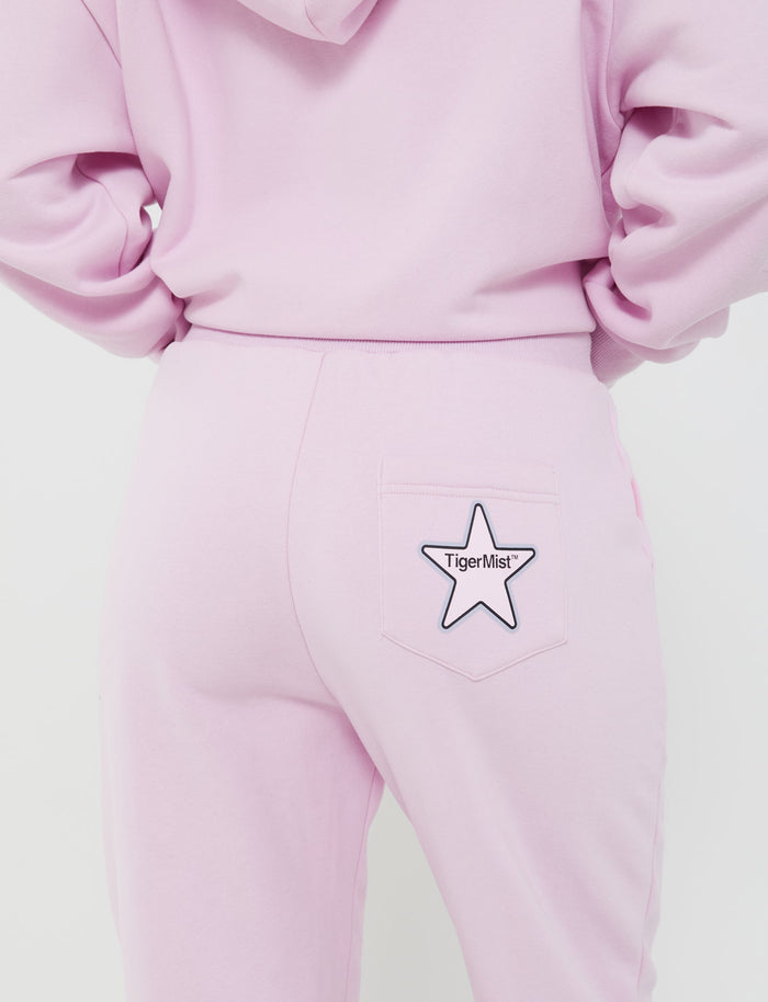 A person is seen from the back wearing the LUMEN PANT - PINK and a matching hoodie. The person has their hands in the hoodie's pockets. There is a star-shaped patch with the text "TigerMist" on the back pocket of the mid-rise waist sweatpants.
