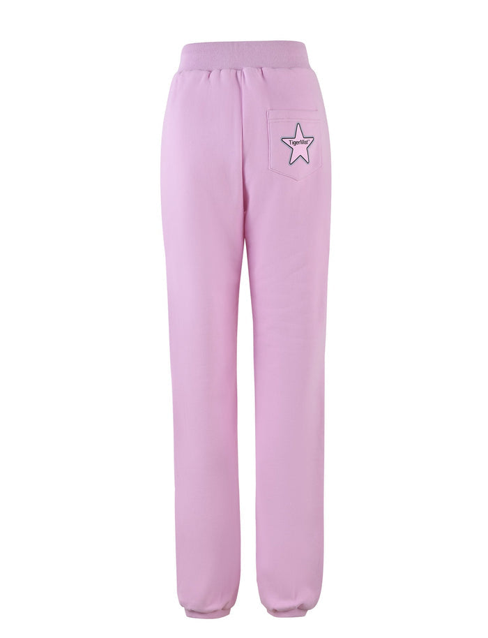 The image displays the TIGERMIST LUMEN PANT in pink, showcasing a pair of relaxed fit, mid-rise loungewear pants with a ribbed waistband and cuffs. A small patch featuring a star and the word "Super" adorns the right back pocket, adding a decorative touch. The oversized trackpant is set against a plain white backdrop.
