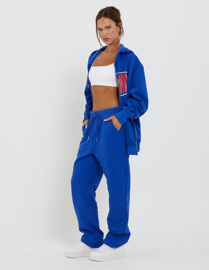 A woman poses confidently against a plain white background. She is wearing the LUMEN ZIP FRONT HOODIE - BLUE from TIGERMIST, accompanied by matching drawstring pants in a royal blue hue. Underneath the zip-up hoodie, she sports a white crop top. Her look is completed with white platform sneakers.
