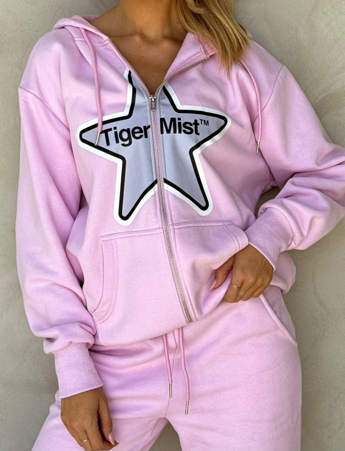 A person is wearing the TIGERMIST Lumen Zip Front Hoodie in pink along with matching sweatpants. The oversized silhouette of the hoodie features a large black and white star graphic with the text "TIGERMIST" inside the star. Made from fleece-lined tracksuiting fabric, the hoodie is zipped partially, and their right hand rests on their hip.