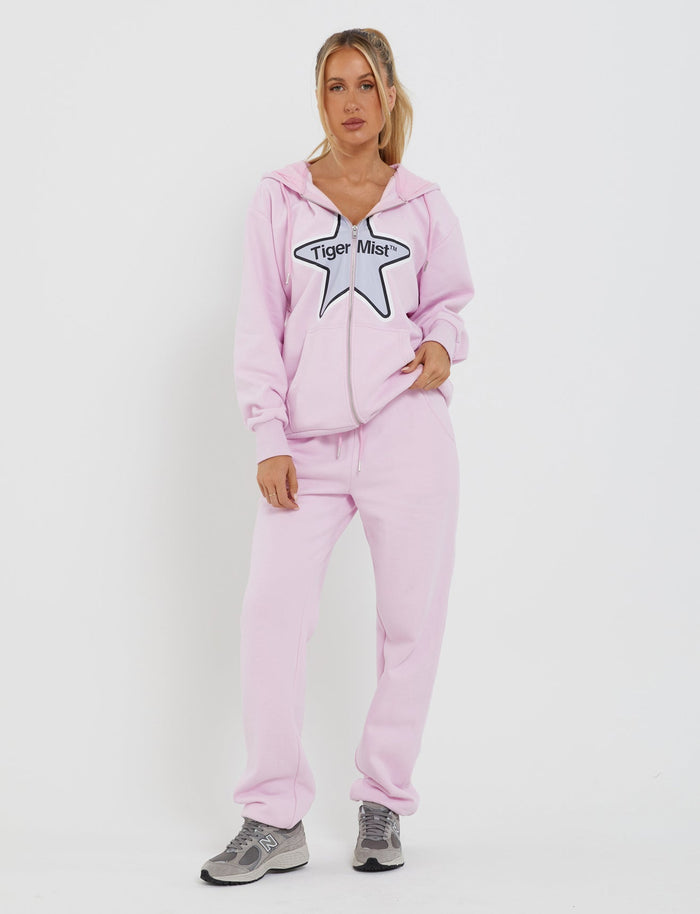 A person stands against a white background, dressed in an oversized silhouette light pink loungewear hoodie and matching sweatpants. The hoodie, named the LUMEN ZIP FRONT HOODIE - PINK from the brand TIGERMIST, is made from fleece-lined tracksuiting fabric and features a star graphic with the text "Tiger Mist" on the front. They have one hand in their pocket and are wearing grey sneakers.