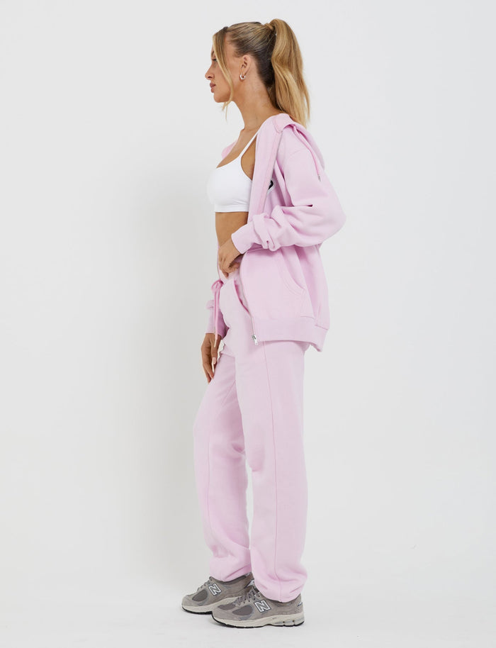 A woman with a ponytail stands in profile against a plain white background. She is wearing an oversized silhouette LUMEN ZIP FRONT HOODIE - PINK by TIGERMIST and matching sweatpants, along with a white sports bra and gray sneakers. One hand is in her pocket as she looks to the side.
