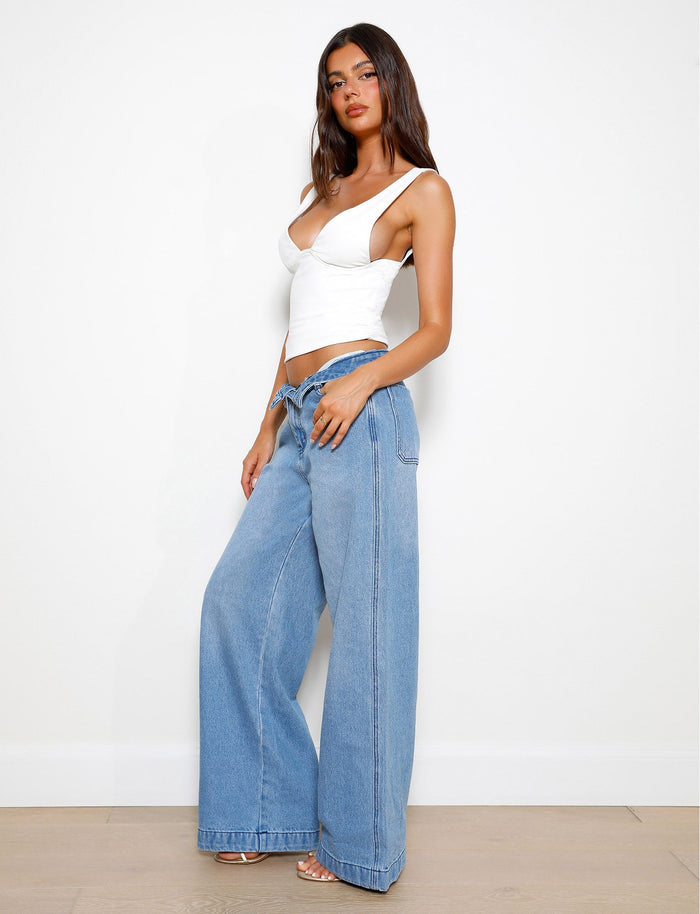 A woman poses against a plain white wall, showcasing her TIGERMIST style. She is wearing the LUNARIA TOP in white and high-waisted, wide-leg blue jeans. With long dark hair, she stands confidently with one hand on her hip and the other hanging by her side, completing the look with neutral-colored sandals.