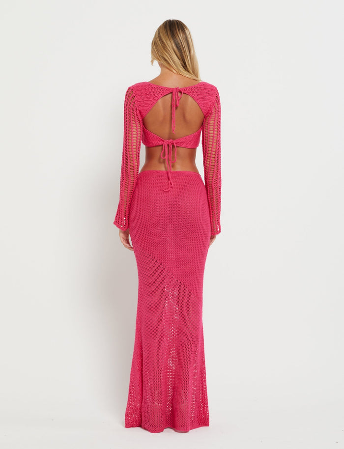 A woman with long blonde hair stands facing away, showcasing the Tiger Mist "MACAULEY SKIRT - HOT PINK." The vibrant pink crochet outfit has a top featuring an open back with tie details, while the form-fitting long skirt extends to the floor. The background is plain white.