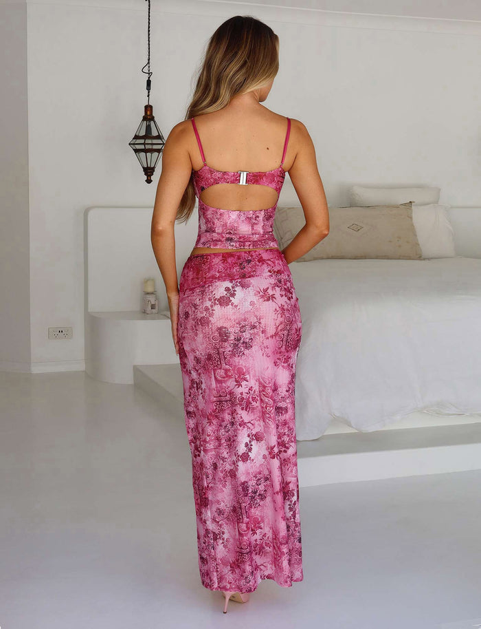 A woman with long hair is standing in a minimalist bedroom, facing away from the camera. She is wearing a pink floral patterned two-piece outfit from TIGERMIST, featuring the MAIZIE SKIRT - MULTI : FLORAL : PINK FLORAL and a matching crop top. The room features a white bed, a modern light fixture, and simple decor.