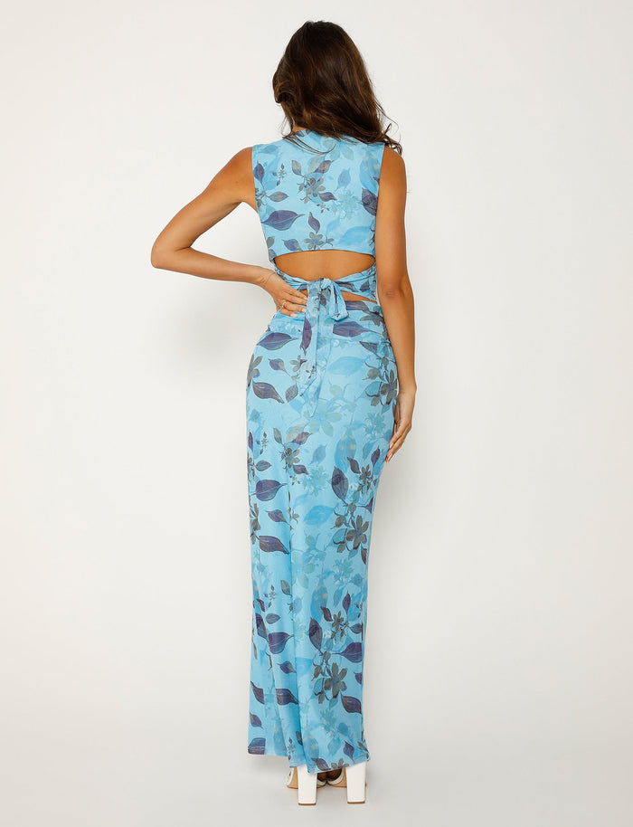 A woman with long wavy hair is facing away from the camera, dressed in a sleeveless Tiger Mist Marietta Skirt - Blue: Watercolour with an open back and a tie detail. The floor-length garment pairs elegantly with her white high heels, as she rests her right hand on her hip.