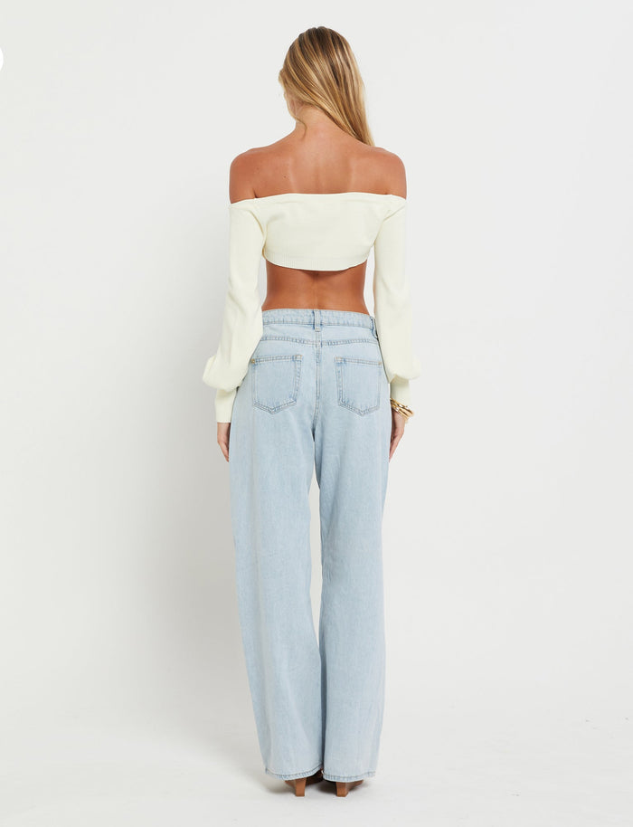 A woman stands facing away from the camera, showcasing her off-the-shoulder long-sleeve MARIYAH Jumper in cream by TIGERMIST and light blue high-waisted jeans. She has long, blonde hair that flows down her back and is standing against a plain white background.