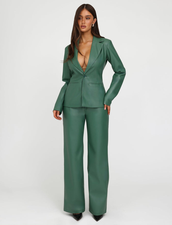 A woman stands against a plain white background, wearing TIGERMIST's deep green MCKAY BLAZER paired with matching leather pants. The fitted blazer features a deep V-neckline and wide lapels, while the pants are wide-legged. She has long, straight hair and wears black pointed-toe shoes.