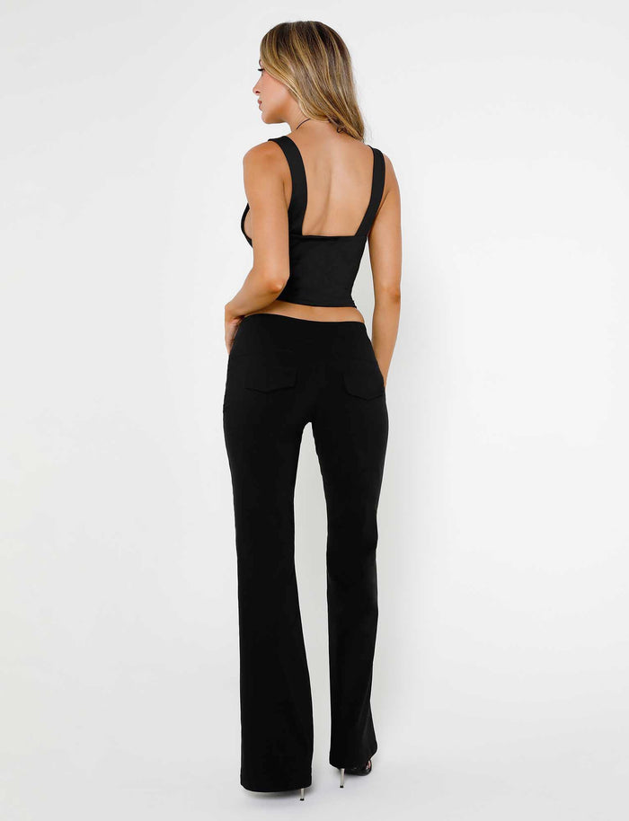A person with long hair is standing and facing away from the camera on a plain background. They are wearing a sleeveless black top with a low back and the MERRI PANT in black from TIGERMIST. They stand on high heels, showcasing a stylish and elegant outfit.