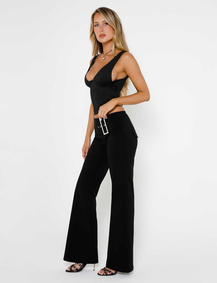 A woman in a black sleeveless top and the MERRI PANT - BLACK from TIGERMIST poses confidently against a white background. She is wearing black high heels and holding a small black clutch with a silver chain. Her long blonde hair cascades over her shoulders, perfectly embodying the chic style of TIGERMIST.