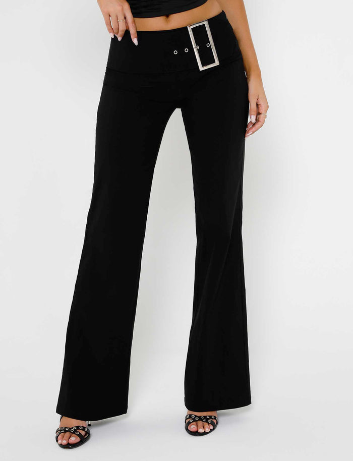 A person is wearing the MERRI PANT - BLACK from TIGERMIST, featuring a high-waisted flare design with a large belt buckle. Their top and face are outside the frame. They are also sporting black strappy high-heeled sandals against a plain white background.