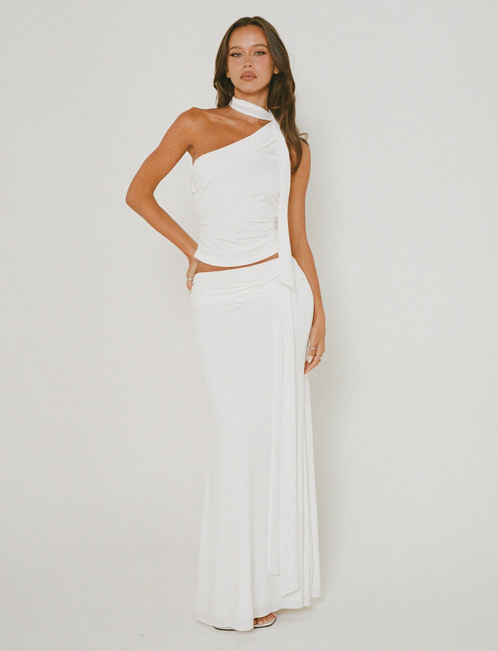 A woman stands against a plain background, wearing the stylish MIST SKIRT - WHITE from TIGERMIST. The skirt showcases an elegant wrap design with draped fabric, paired perfectly to highlight its chic one-shoulder style. She poses with one hand on her hip and the other resting by her side.