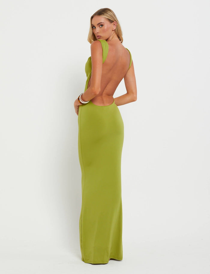 A person with long blonde hair is posing in the MORANA DRESS - GREEN : PASTEL GREEN by TIGERMIST. They are turned slightly to the side, showcasing the backless, floor-length design of the dress. The background is plain white, devoid of any additional objects or scenery.