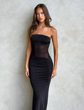 A woman with long, wavy brown hair stands against a textured light grey background, wearing a form-fitting MORGAN DRESS - BLACK by TIGERMIST. She faces the camera with her arms relaxed at her sides. The dress features semi-sheer panels around the waist.