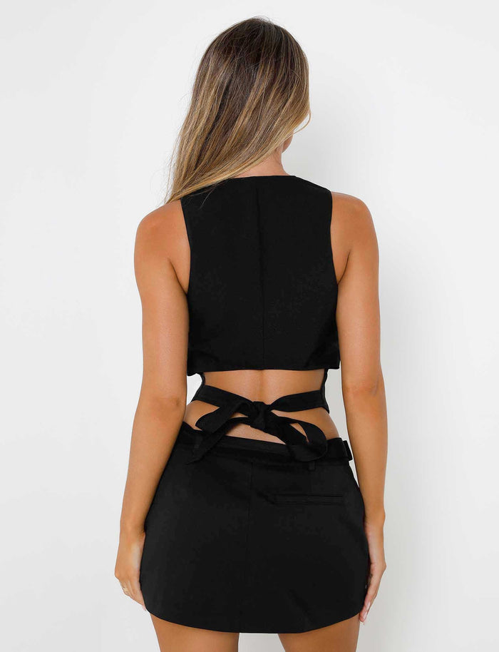 A person with long hair is shown from the back, wearing a sleek, black sleeveless top that ties at the waist and a matching NADINE SKORT - BLACK from TIGERMIST against a plain white background.